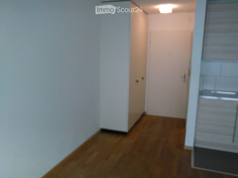 Empty room with wooden floor, white walls, and an entrance leading to another room with an appliance.