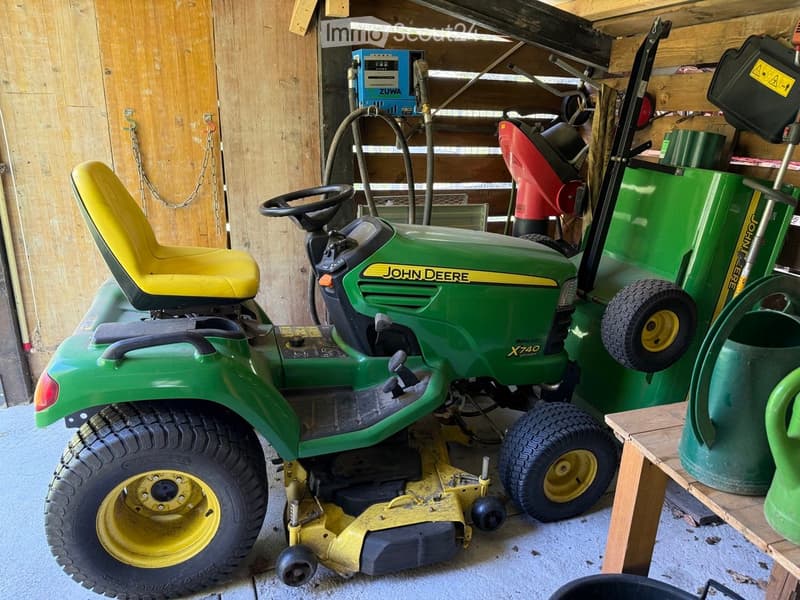 A green John Deere lawn mower with a yellow seat is parked inside a wooden shed, alongside another lawn mower and a small table holding a green can.