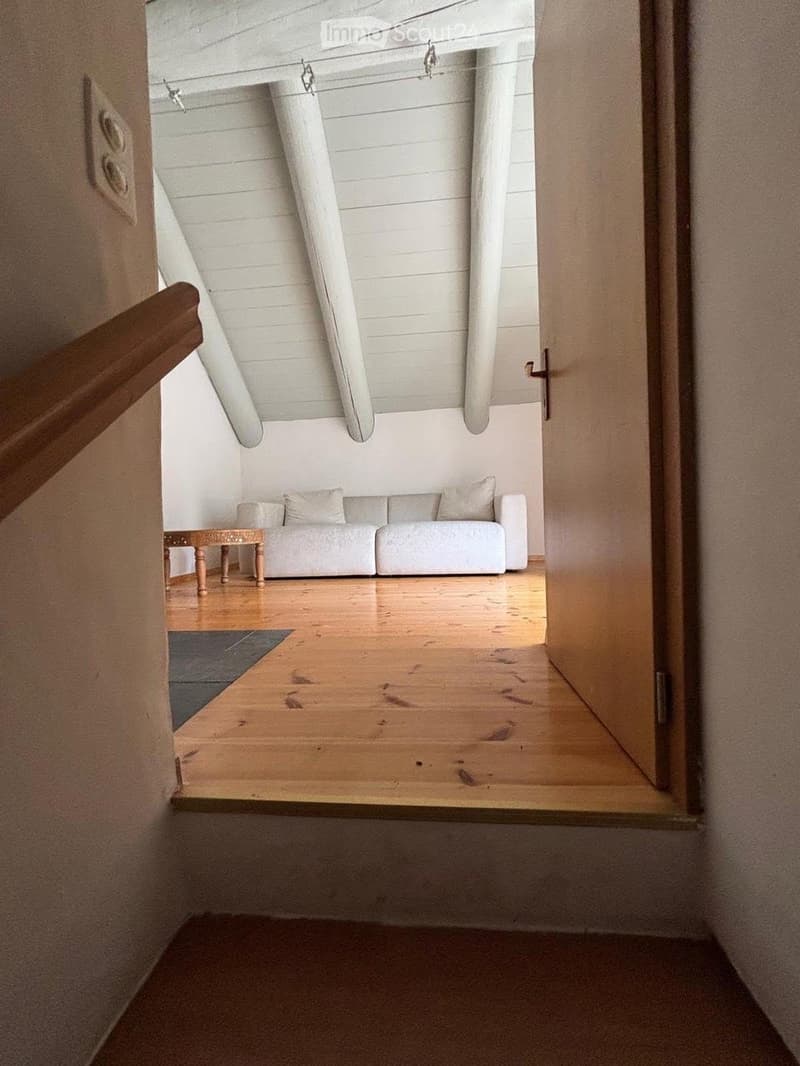 A modern living room with a raised wooden floor, white walls, and exposed wooden beams. Features a white sofa with pillows, a small wooden table, and a gray rug.