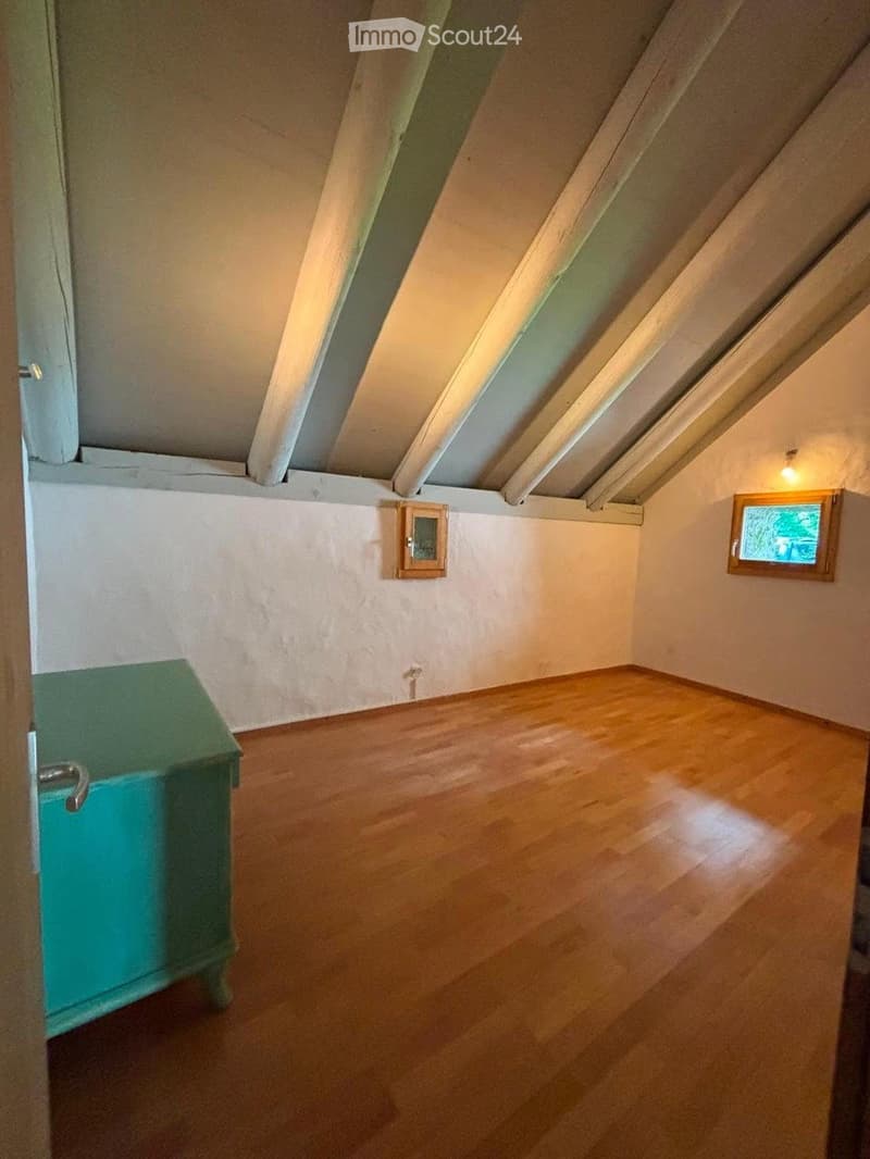 Empty room with wooden flooring, slanted ceiling beams, and two small windows. A small green cabinet is visible on the left side.