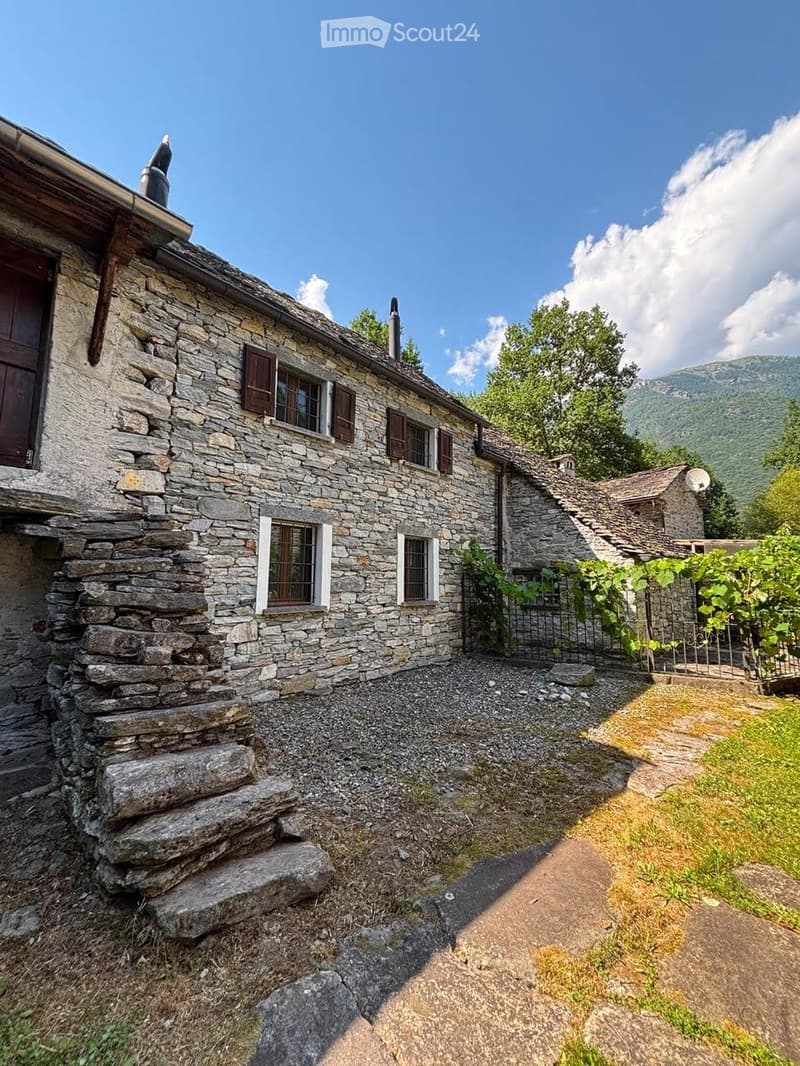A rustic stone house with a sloped roof, wooden shutters, and a small garden area with vines and a gravel pathway.
