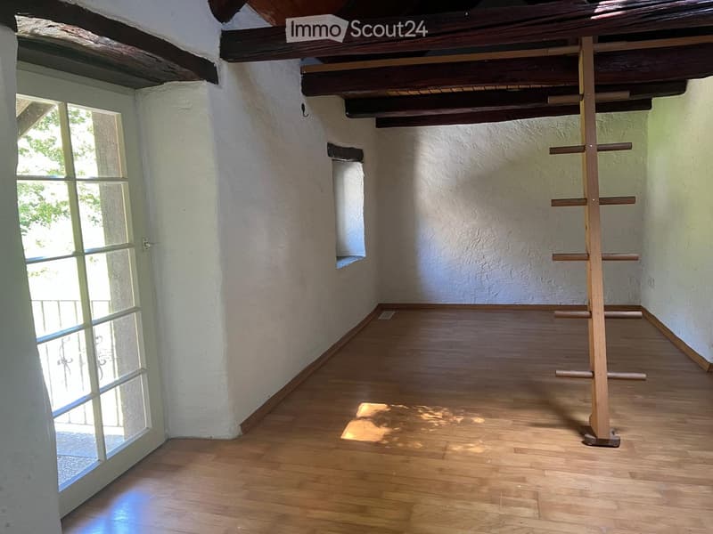Empty room with wooden flooring, white walls, and exposed wooden beams on the ceiling. A large window with glass panels allows natural light to enter, and a wooden drying rack stands in the corner.