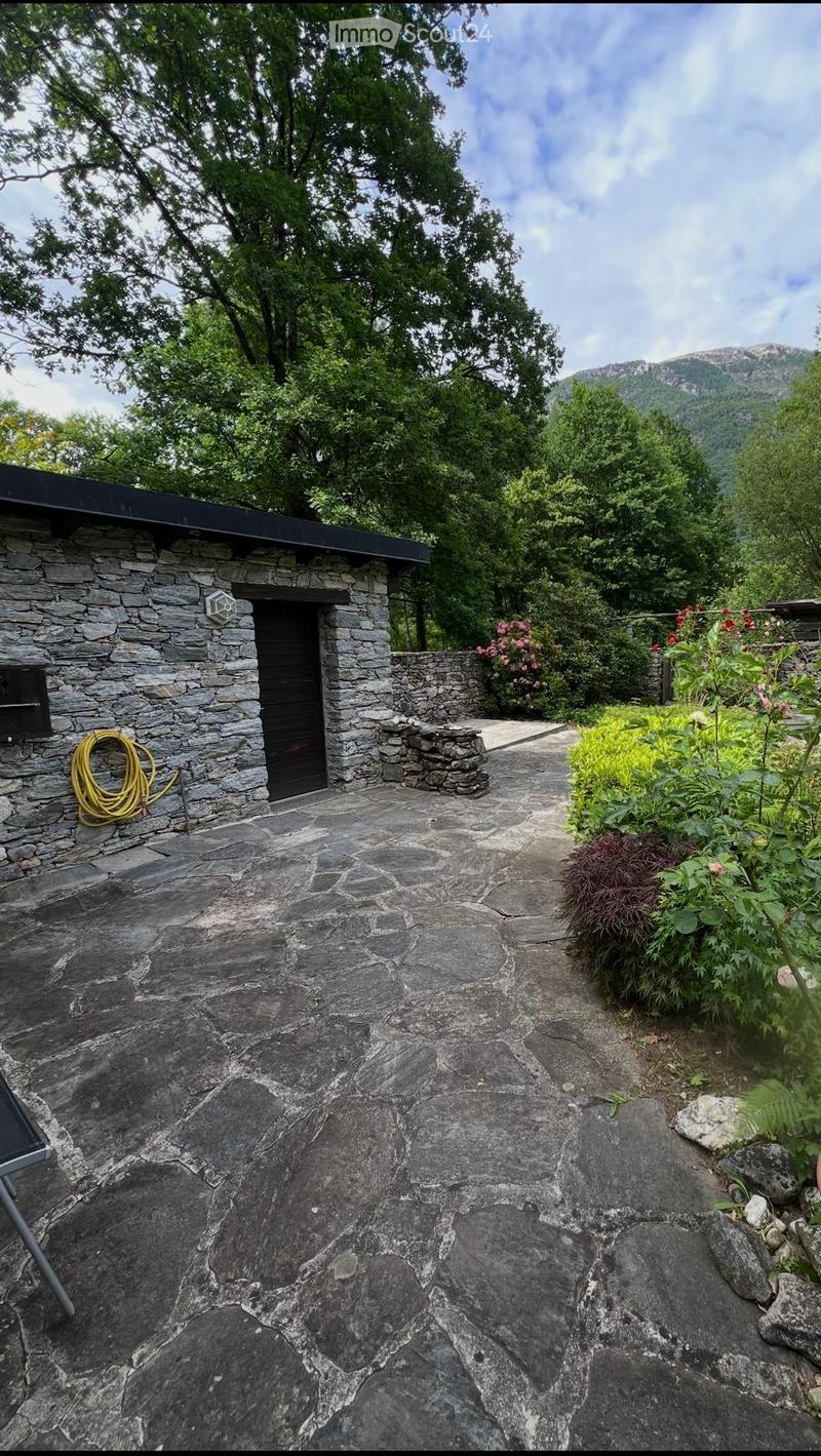 Stone pathway leads to a small stone building with a black roof, surrounded by lush greenery and a mountainous backdrop.