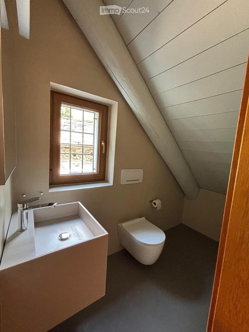 Compact bathroom with a modern sink, sleek faucet, and toilet, featuring a slanted wooden ceiling and a small window with wooden frame.