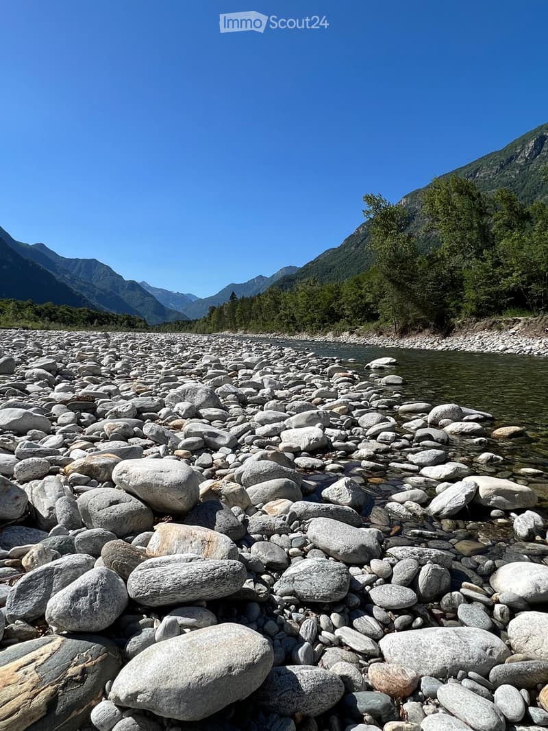 A riverbank with large rocks and a calm river flowing through a mountainous landscape under a clear blue sky.