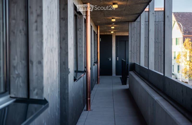 A modern corridor with wooden walls, copper pipes, and a concrete floor. The corridor is illuminated by a series of small lights on the ceiling.