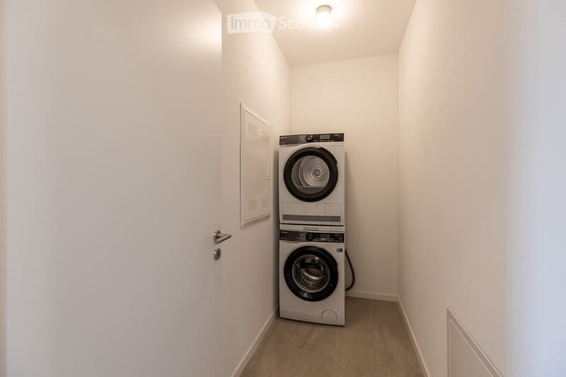 A small, empty room with white walls and wooden flooring. It contains a stack of white washing machines and a white door.