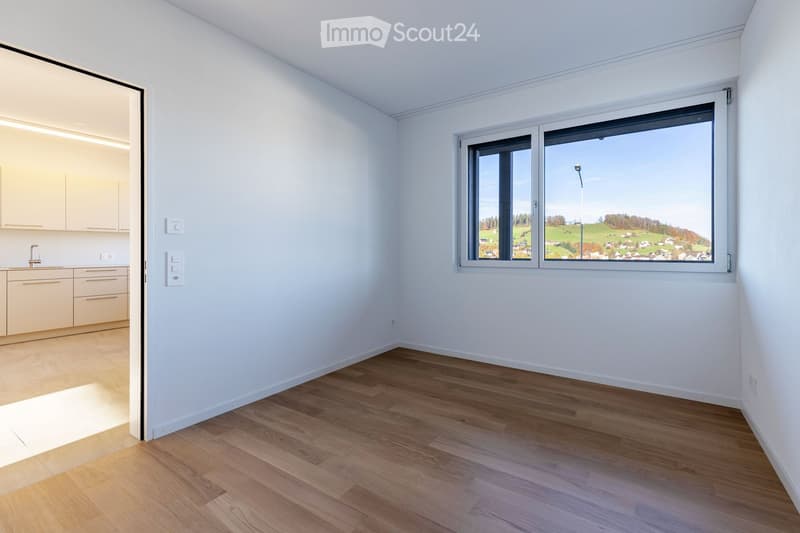An empty room with white walls, wooden floors, and a large window offering a view of a landscape. The room is devoid of furniture.