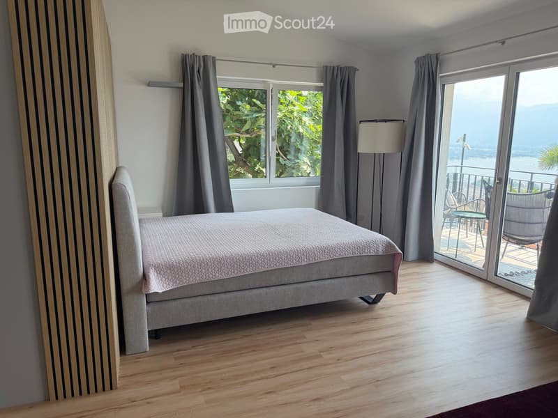 Modern bedroom with wooden flooring, grey upholstered bed, large window with curtains, and sliding glass door leading to a balcony with outdoor furniture and scenic view.