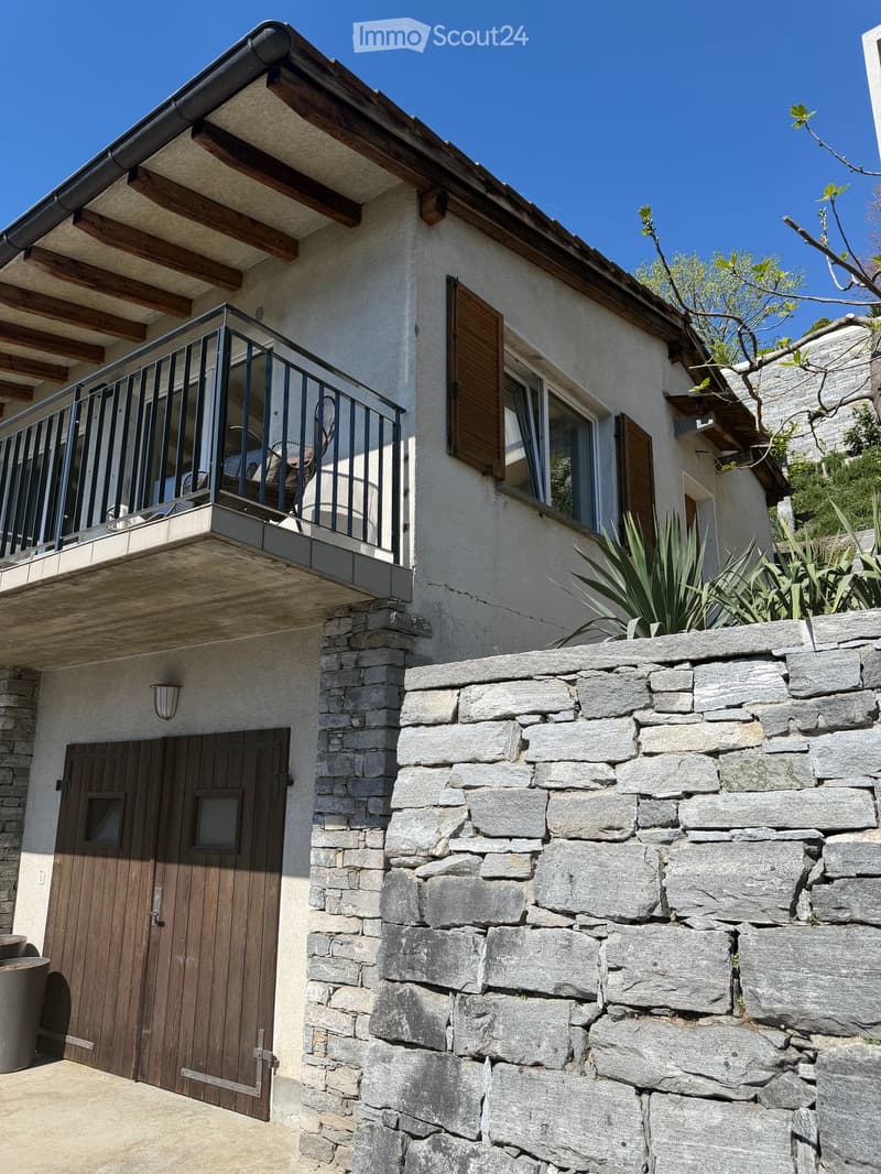 Two-story house with a balcony, wooden shutters, stone wall, and a wooden garage door.