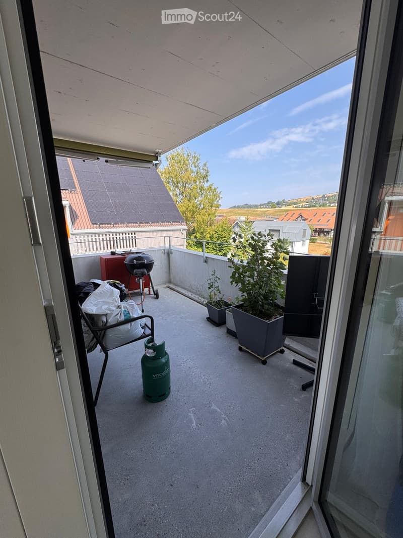 Balcony with a concrete floor, a chair, a grill, potted plants, and a gas cylinder. The balcony has a view of the surrounding area with trees and buildings.