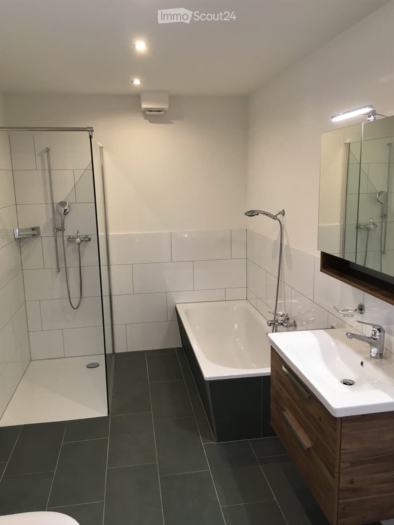 Modern bathroom with a bathtub, sink, mirror, and shower area. Features dark tiled flooring and white tiled walls.