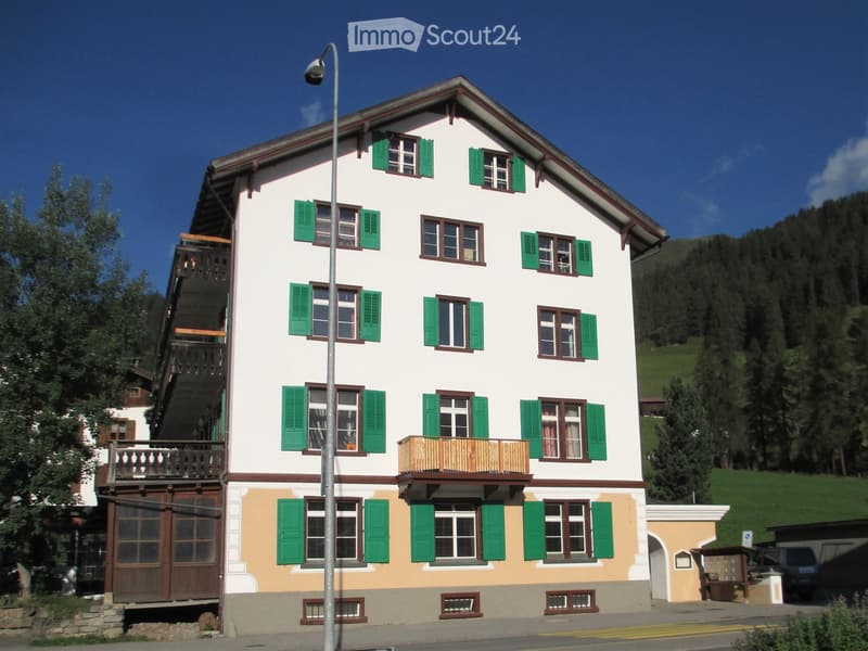 A two-story house with white walls, green shutters, and wooden balconies, situated in a scenic, mountainous area.
