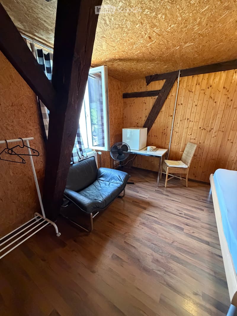 Cozy attic room with wooden walls and floors, featuring a black leather sofa, a small table with a fan and refrigerator, a wicker chair, and a clothes hanger near the window.