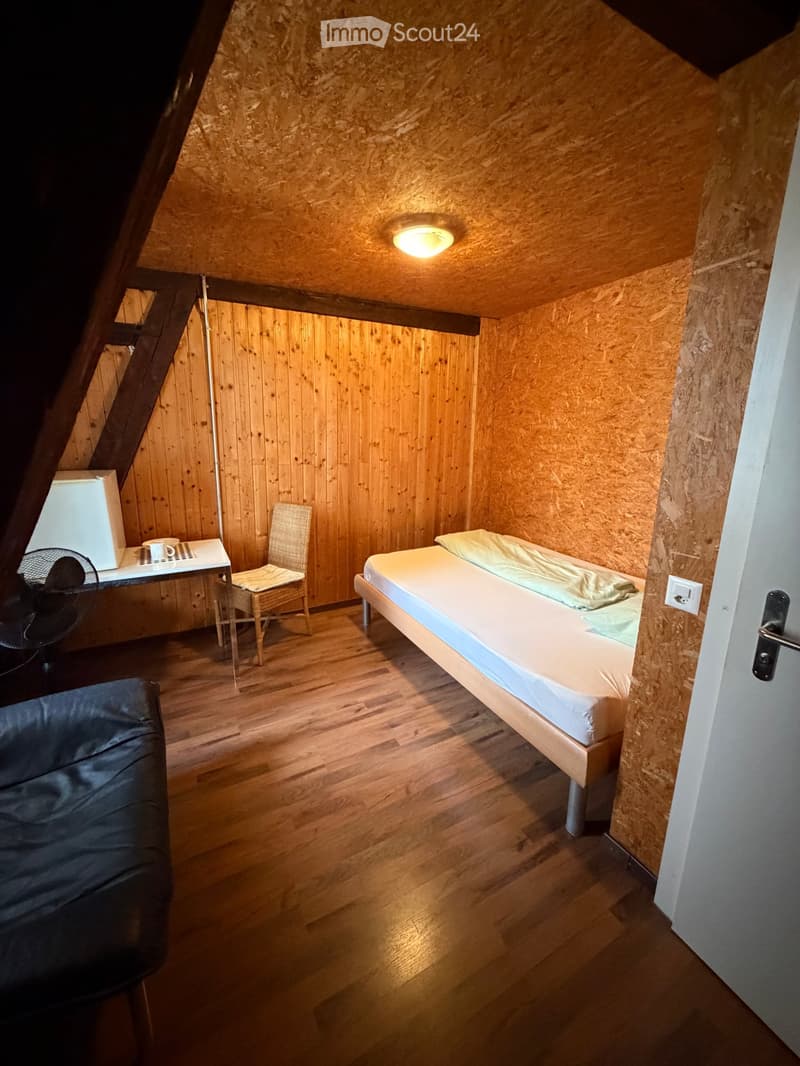 Cozy attic bedroom with wooden walls and floor, featuring a single bed, a small table with a fan and cup, a wicker chair, and a black leather ottoman.
