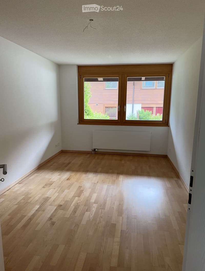 Empty room with wooden flooring, white walls, and large windows with wooden frames. A radiator is mounted on the wall below the windows.