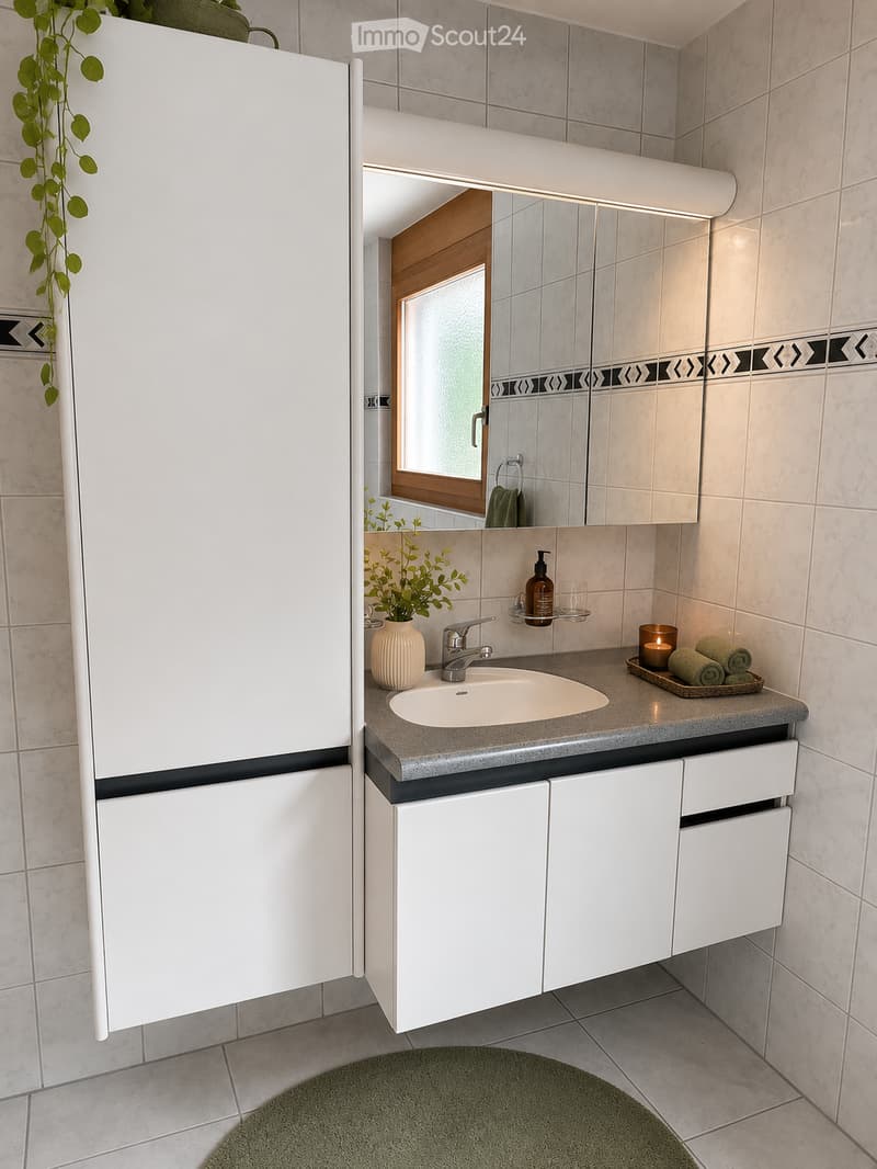 Modern bathroom with a white sink, chrome faucet, large mirror, white cabinets, decorative vase with plant, candle, and folded towels on the counter.