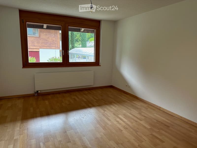 Empty room with wooden flooring, white walls, large window with wooden frame, radiator, and visible light fixture.