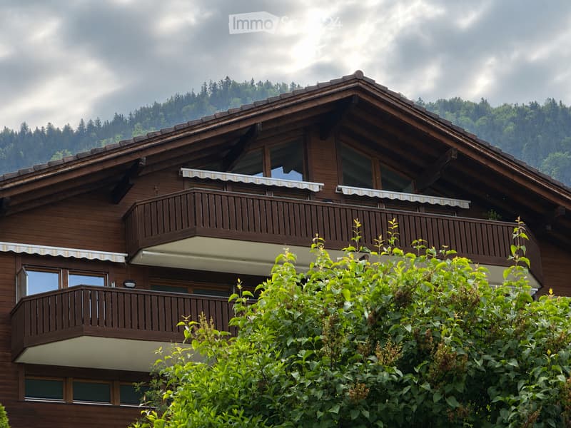 A two-story wooden house with balconies, large windows, and striped awnings, surrounded by greenery and trees.