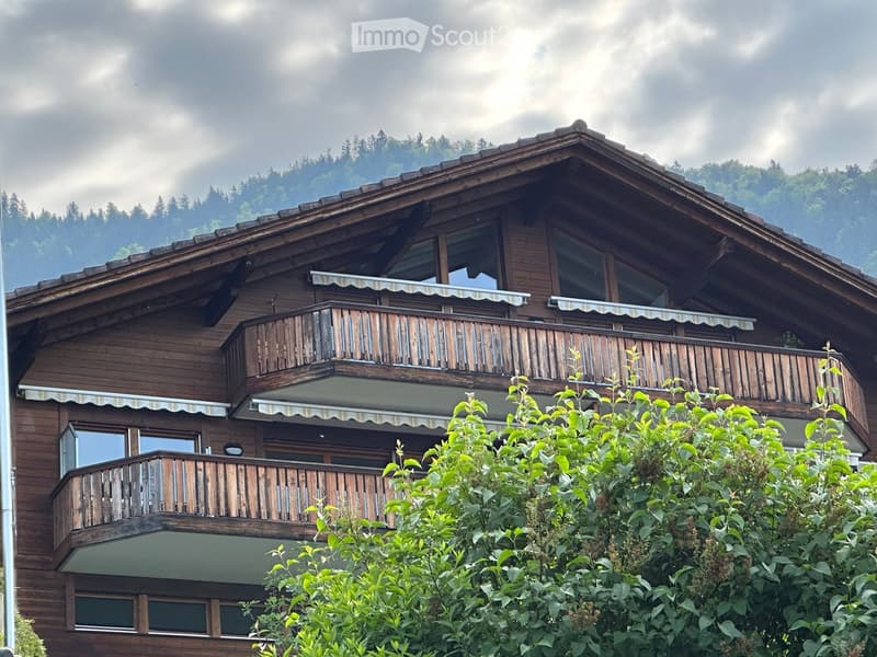 A two-story wooden house with balconies, large windows, and a sloped roof, surrounded by greenery and trees.