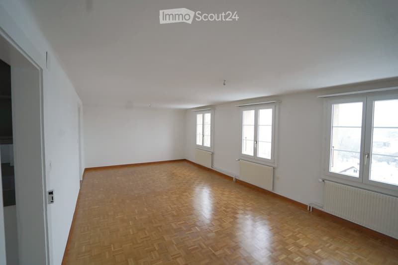 Empty room with wooden flooring, white walls, and multiple windows allowing natural light. Radiators are visible along the walls.
