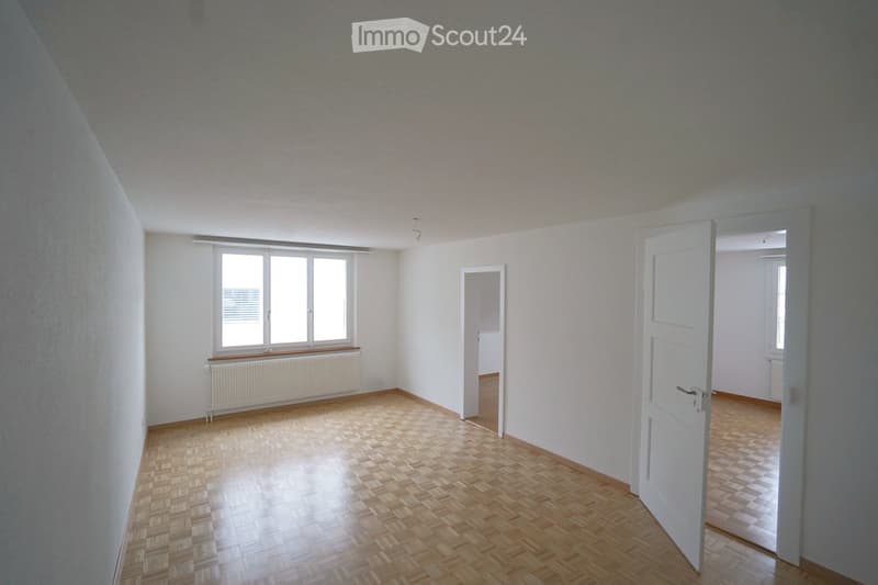 Empty room with wooden flooring, white walls, large window with radiator below, and two open doors leading to adjacent spaces.