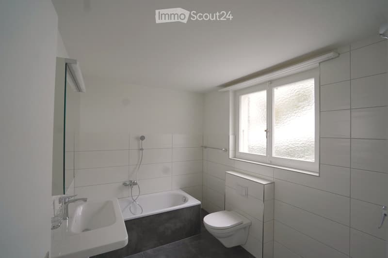 Modern bathroom with white tiles, a bathtub, sink, toilet, and a large window providing natural light.