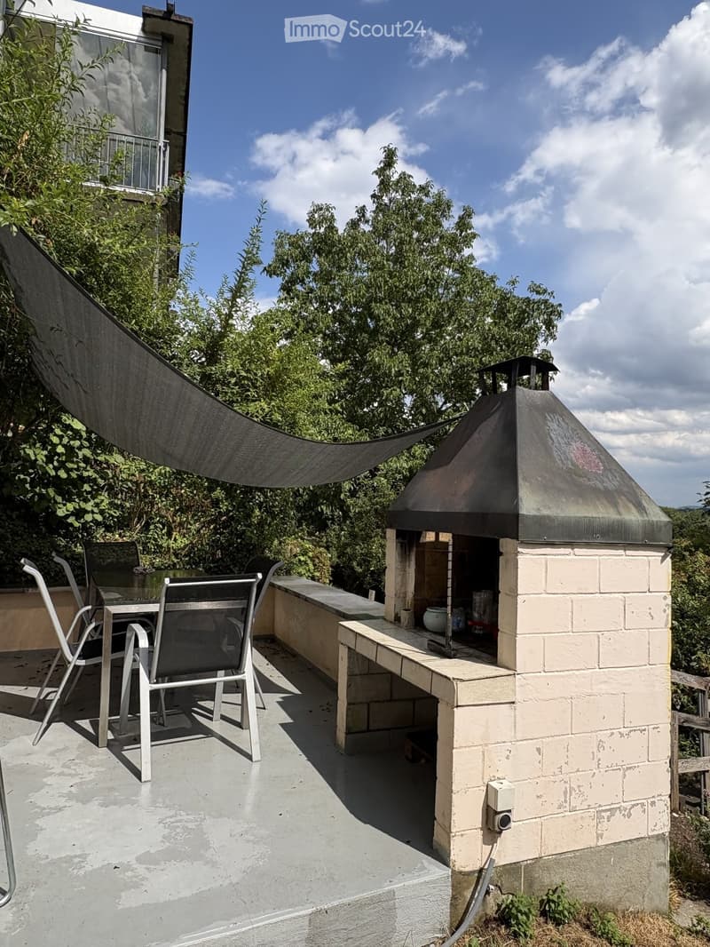 Balcony with a table, chairs, and a brick fireplace with a metal hood, surrounded by greenery and a clear sky.