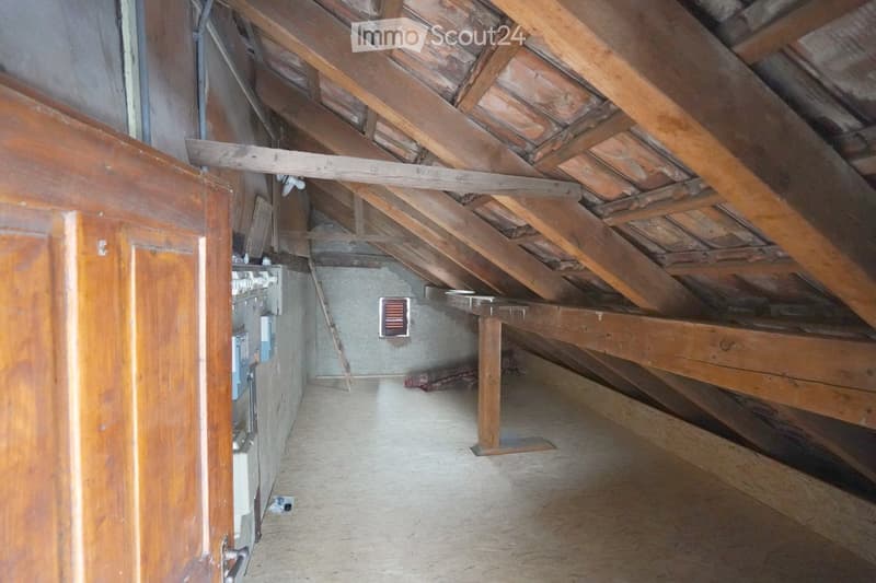An attic space with exposed wooden beams and rafters, concrete floor, and minimal furnishings. The area includes a small wooden platform and some electrical components on the wall.