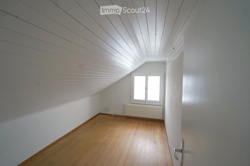 Empty attic room with wooden flooring, white walls, and a slanted ceiling. Features a window with two panes and a radiator below it.