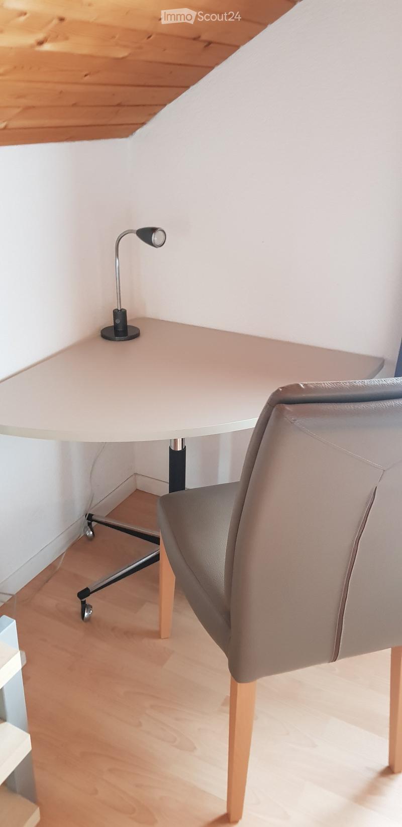 A modern, minimalist room with a light-colored wooden floor, a white desk, a grey upholstered chair, and a sleek black desk lamp.
