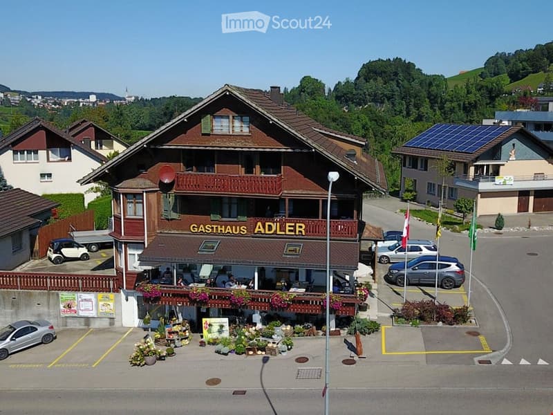 A traditional Swiss restaurant named "Gasthaus Adler" with a wooden facade, balcony, and outdoor seating area. The building has multiple stories with a pitched roof and is surrounded by parked cars and greenery.