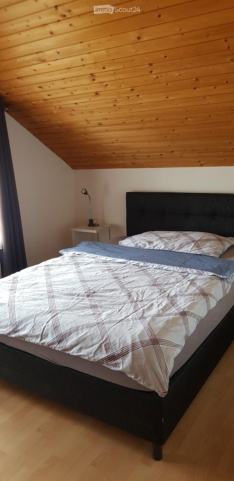 Cozy bedroom with wooden walls and floor, featuring a neatly made bed with a plaid duvet and pillows, a nightstand with a lamp, and a sleek black headboard.