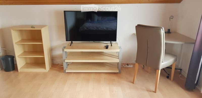 A small, furnished room with a wooden floor, a television on a stand, a chair, a small table, and a shelf.