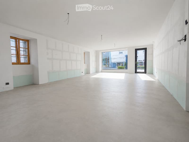 Empty room with white walls, light grey carpeted floor, and large windows allowing natural light. Features include a wooden-framed window, a glass door leading outside, and a partially painted wall in light blue.