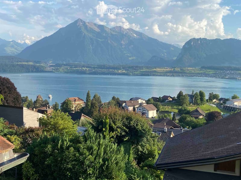 Panoramic view of a lake and surrounding mountains from a garden area, with houses and trees in the foreground.