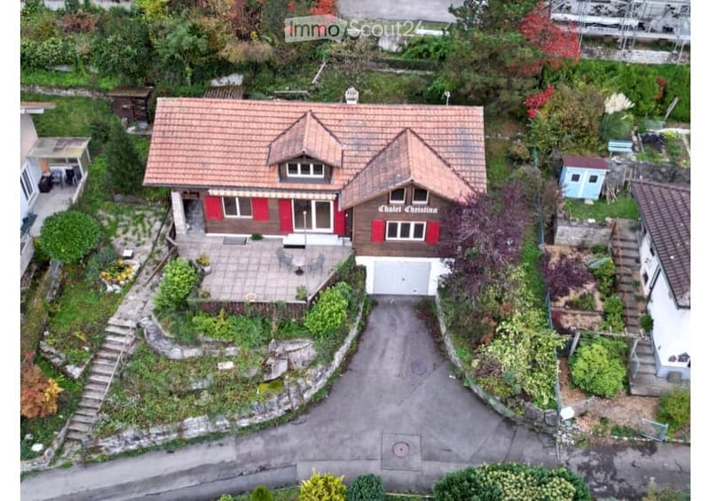 A two-story house with a red-tiled roof and red shutters, surrounded by a well-maintained garden and driveway. The house features a garage and a small patio area with outdoor furniture.