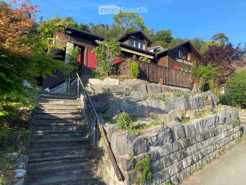 A multi-story wooden house with a stone staircase leading up to it, surrounded by greenery and trees.