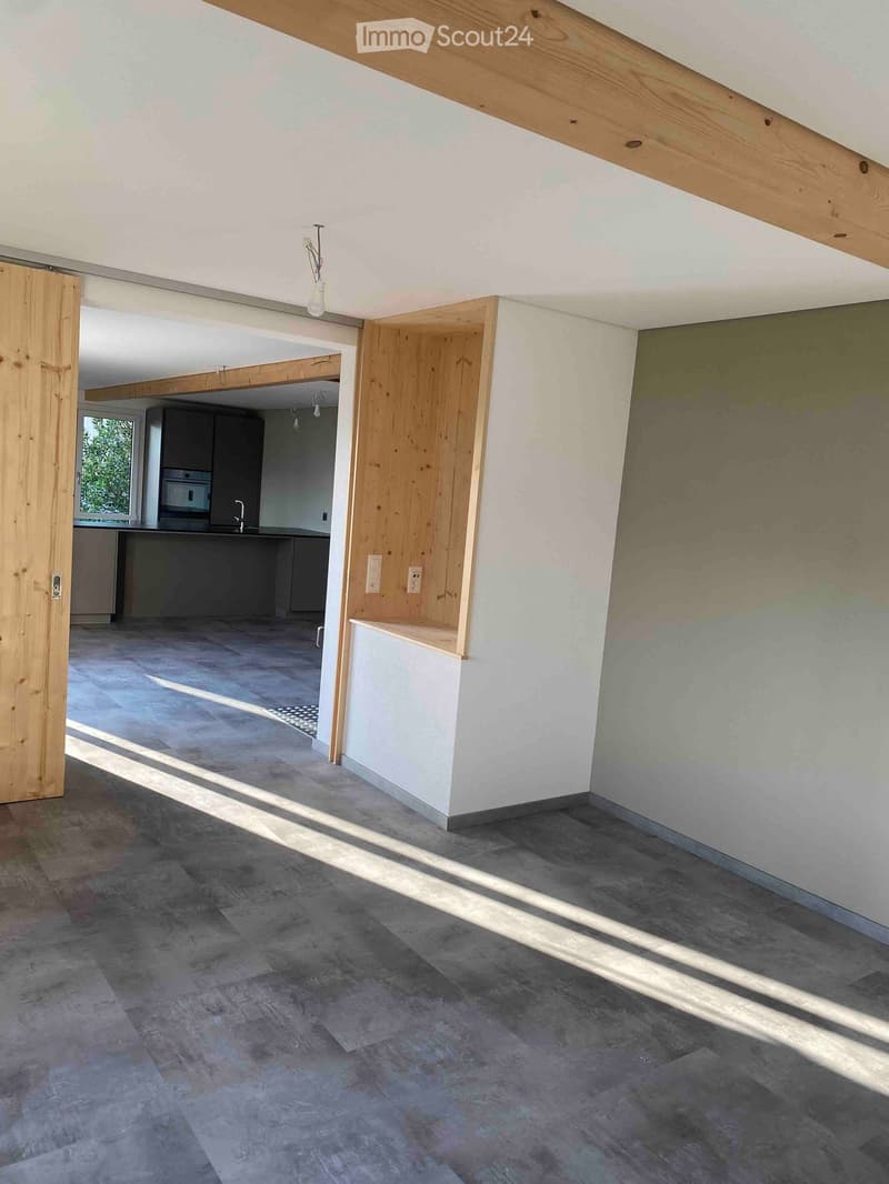 Empty room with grey tiled floor, white walls, wooden door frame, and visible kitchen area in the background.