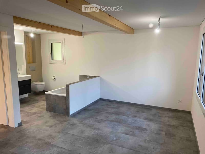 Modern bathroom with a freestanding bathtub, sink, toilet, and mirror. Exposed wooden beams on the ceiling and large windows provide natural light.