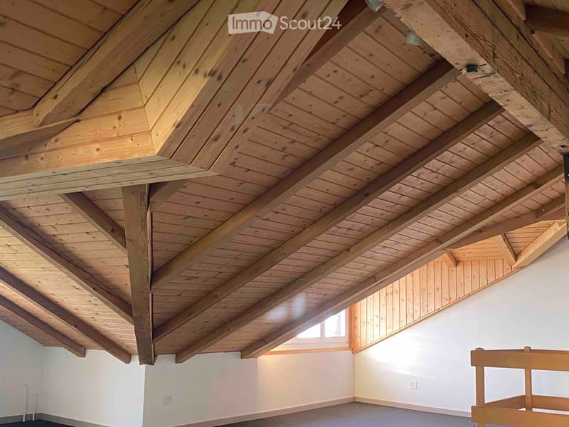 An empty room with exposed wooden beams and a slanted wooden ceiling, white walls, and a small window letting in natural light.