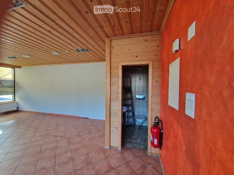 Empty room with wooden ceiling, tiled floor, and a doorway leading to a small bathroom. The walls are painted in neutral tones, and there is a fire extinguisher mounted on the wall near the doorway.