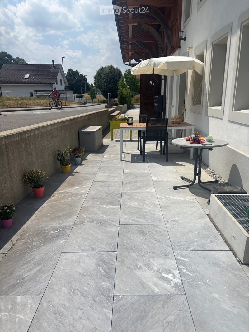 Balcony with concrete floor, outdoor furniture including a table, chairs, umbrella, and flower pots.