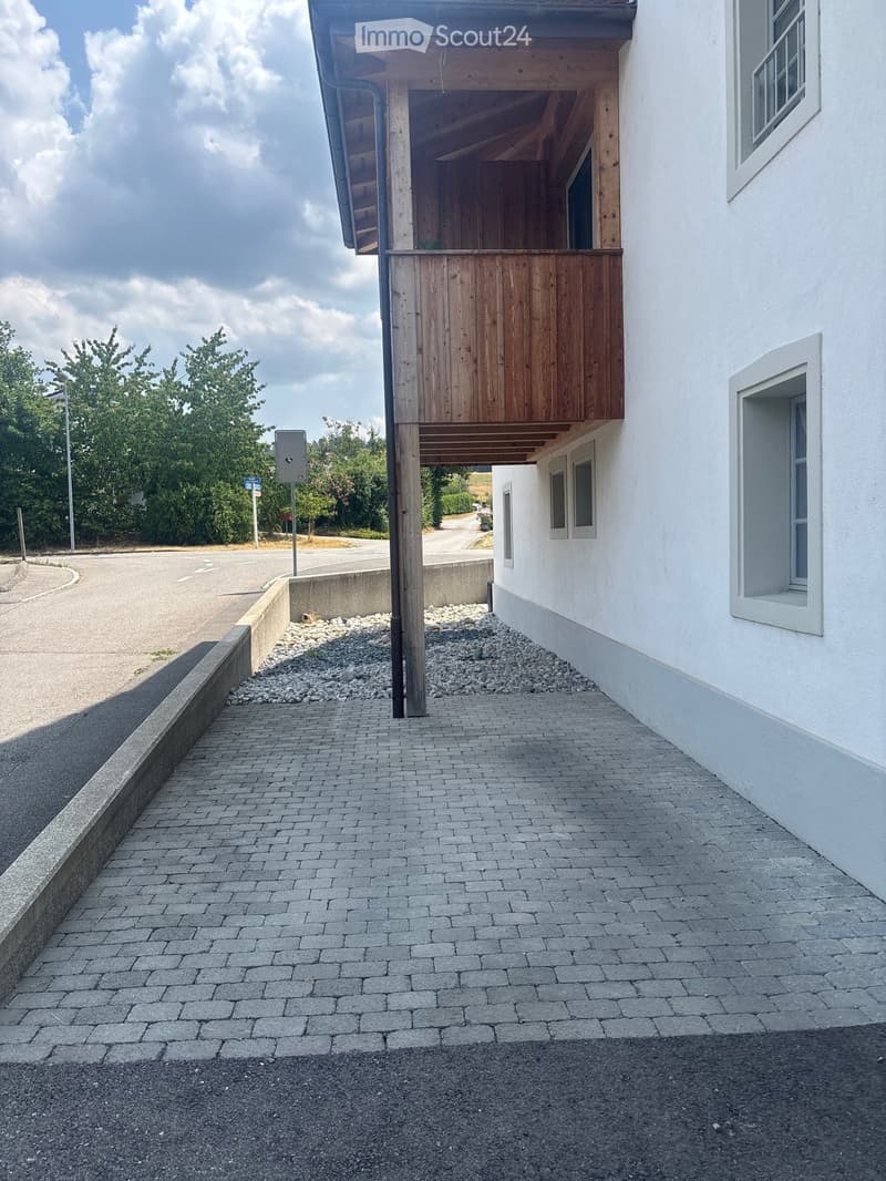 Modern two-story building with a wooden balcony and white exterior walls, situated on a paved pathway with a gravel border.