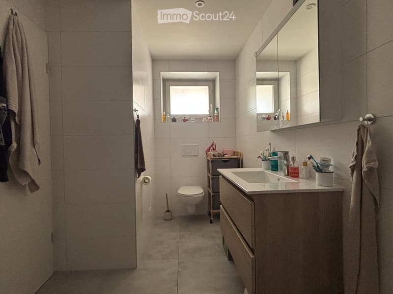 Modern bathroom with a white sink, mirror, toilet, and various toiletries. Light-colored tiles and a window provide natural light.