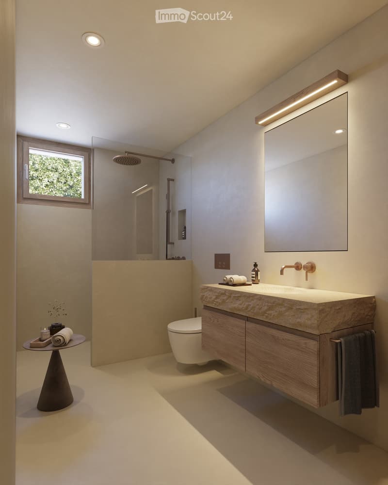 Modern bathroom with a large sink, mirror, toilet, and a small table with towels and a bottle. Features a shower area with a glass enclosure and a window letting in natural light.