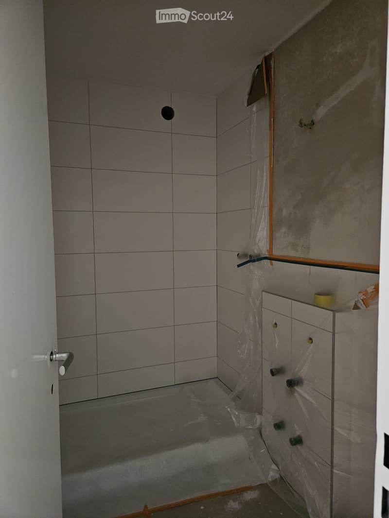 Empty bathroom with tiled walls, a door, and protective plastic sheeting on the floor and part of the wall.