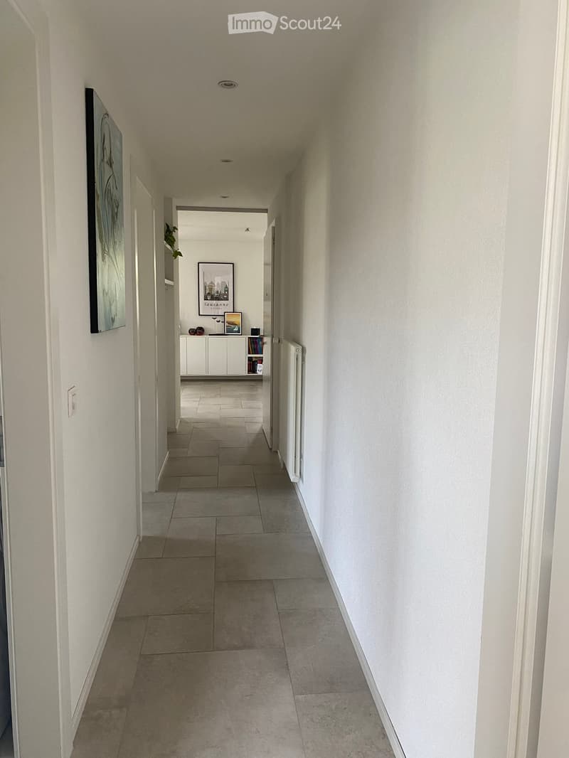 A clean, well-lit hallway with tiled flooring and white walls. A small cabinet with books and decorative items is visible at the end of the corridor.