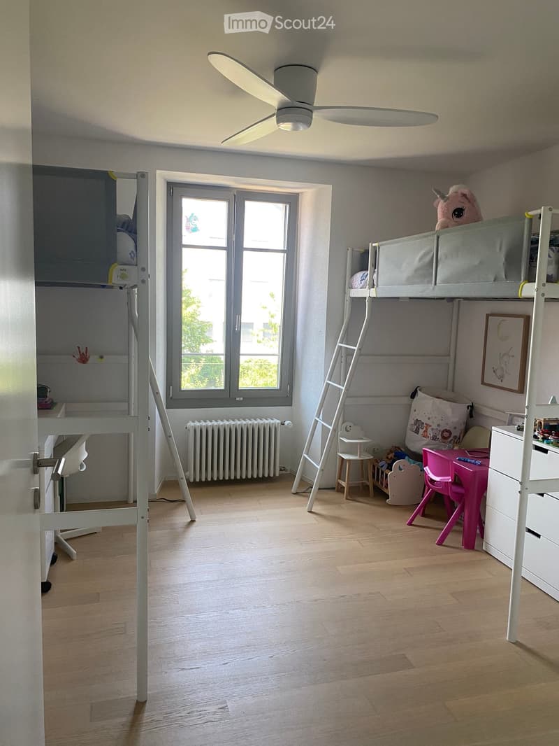 Brightly lit bedroom with wooden flooring, bunk bed, pink chair, small table, and various toys.