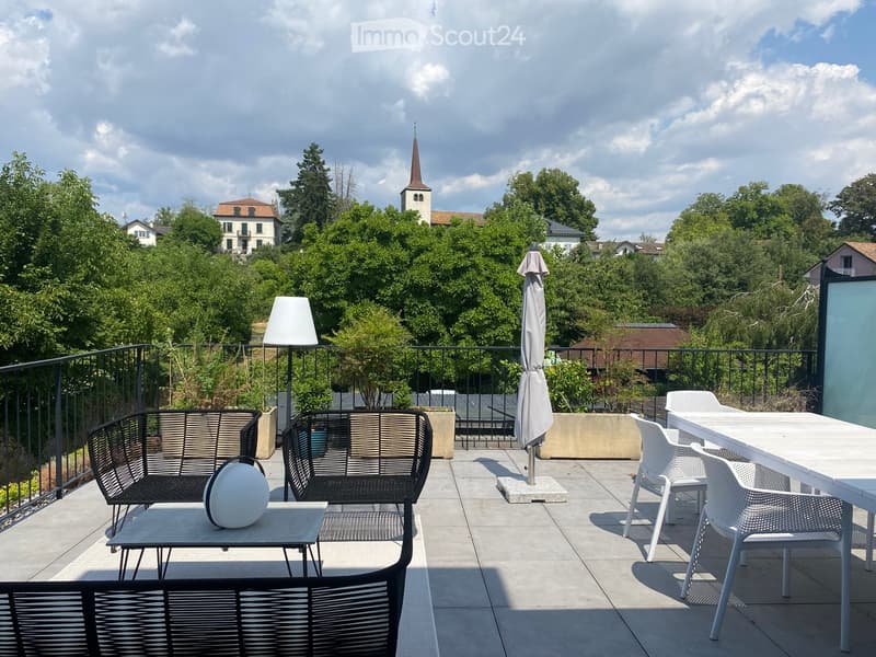 Balcony with outdoor furniture including a sofa, table, chairs, and an umbrella, overlooking a scenic view of trees and a church spire.